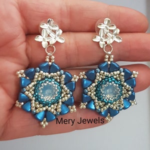 May include: A pair of blue and silver star-shaped earrings with a clear crystal center. The earrings are hanging from silver flower-shaped posts. The earrings are labeled "Mery Jewels".