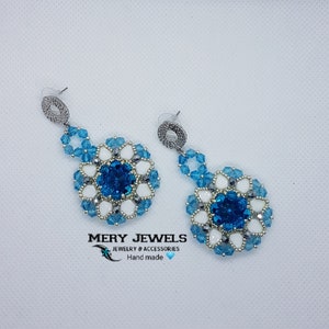 Può includere: Un paio di orecchini pendenti con un design di perline blu e bianche. Gli orecchini presentano un grande cristallo blu circondato da perline bianche e perline blu più piccole. Gli orecchini sono argentati e hanno una semplice chiusura a gancio. Mery Jewels Gioielli & Accessori Fatto a mano.