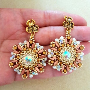 Może przedstawiać: Para złotych i różowo-złotych kolczyków wiszących z koralikowym wzorem płatka śniegu i przezroczystym, tęczowym kryształem w środku.