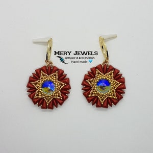 以下が含まれることがあります： 青色の虹色のセンターを持つ、赤と金の星型のイヤリングのペア。イヤリングは手作りで、金色のフック留め具が付いています。イヤリングには「MERY JEWELS ジュエリー＆アクセサリー 手作り」とラベルが貼られています。