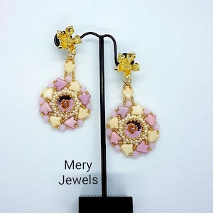 Può includere: Un paio di orecchini a forma di fiore con perline rosa e crema e dettagli dorati. Gli orecchini sono appesi a un supporto nero. Gli orecchini hanno un centro rosa. Il testo "Mery Jewels" è visibile sotto gli orecchini.