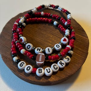May include: Three stacked beaded bracelets featuring red, white, and black seed beads. One bracelet displays the text OHIO with red heart beads, another features a brown football charm, and the third displays the text GO BUCKS in white beads.