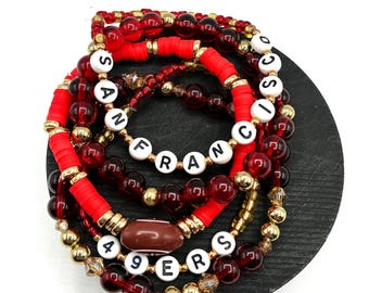 May include: A stack of beaded bracelets in red, gold, and clear beads. Some bracelets spell out "SAN FRANCISCO 49ERS" with white letter beads. The bracelets are on a black, round surface.