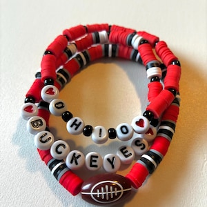 May include: A set of three stacked bracelets made of red, black, white, and gray clay disc beads. One bracelet features white letter beads spelling OHIO and BUCKEYES with red heart accents. A brown football-shaped bead is centered on one strand.