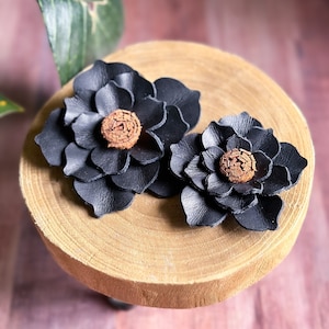 Pode incluir: Dois clipes de cabelo de couro preto em forma de flor com centros marrons. Os clipes s&atilde;o exibidos em uma superf&iacute;cie de madeira.