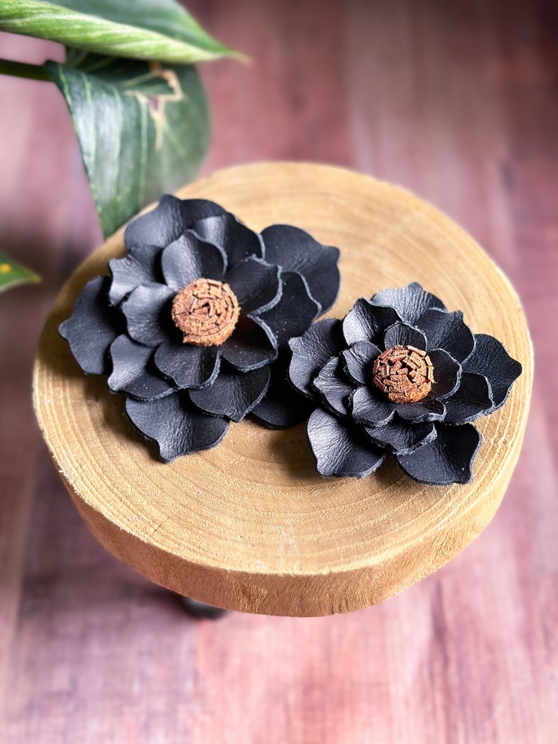 Pode incluir: Dois clipes de cabelo de couro preto em forma de flor com centros marrons. Os clipes s&atilde;o exibidos em uma superf&iacute;cie de madeira.