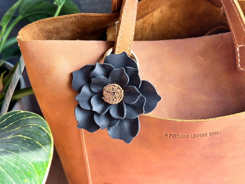 Pode incluir: Uma bolsa tote de couro marrom com um charme de flor de couro preto preso &agrave; al&ccedil;a. A bolsa tem o texto "Portland Leather Goods" gravado na parte inferior.