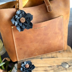 Pode incluir: Uma bolsa tote de couro marrom com um enfeite de flor de couro preto. A bolsa tem um bolso frontal com o texto "Portland Leather Goods" em relevo.