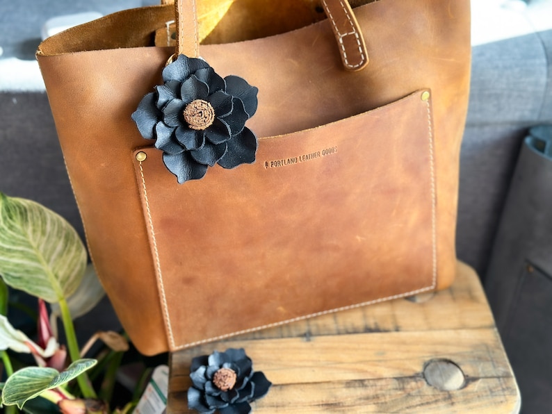 Pode incluir: Uma bolsa tote de couro marrom com um enfeite de flor de couro preto. A bolsa tem um bolso frontal com o texto "Portland Leather Goods" em relevo.