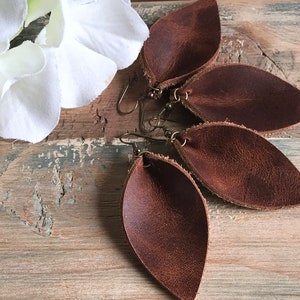 May include: A pair of brown leather earrings with a leaf shape. The earrings are hanging from a wooden surface.