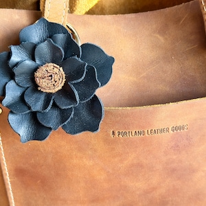 Pode incluir: Um amuleto de flor de couro preto preso a uma bolsa de couro marrom com o texto "PORTLAND LEATHER GOODS" gravado na bolsa.
