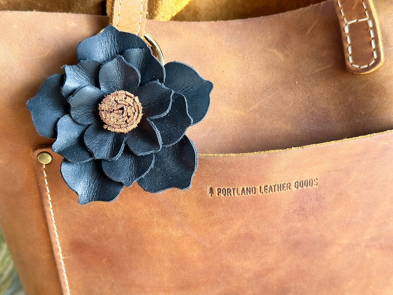 Pode incluir: Um amuleto de flor de couro preto preso a uma bolsa de couro marrom com o texto "PORTLAND LEATHER GOODS" gravado na bolsa.