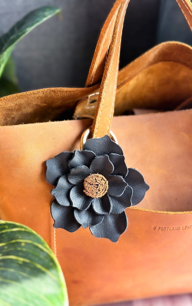 Pode incluir: Uma bolsa tote de couro marrom com um charme de flor de couro preto preso &agrave; al&ccedil;a. A flor tem um centro marrom e a bolsa tem o texto "Portland Leather" gravado na parte inferior.