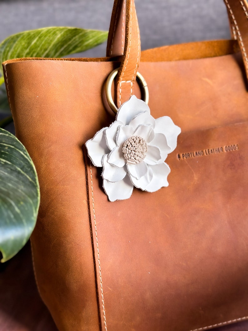 Pode incluir: Uma bolsa tote de couro marrom com um charme de flor de couro branco preso &agrave; al&ccedil;a. A bolsa tem o texto "Portland Leather Goods" impresso no lado.