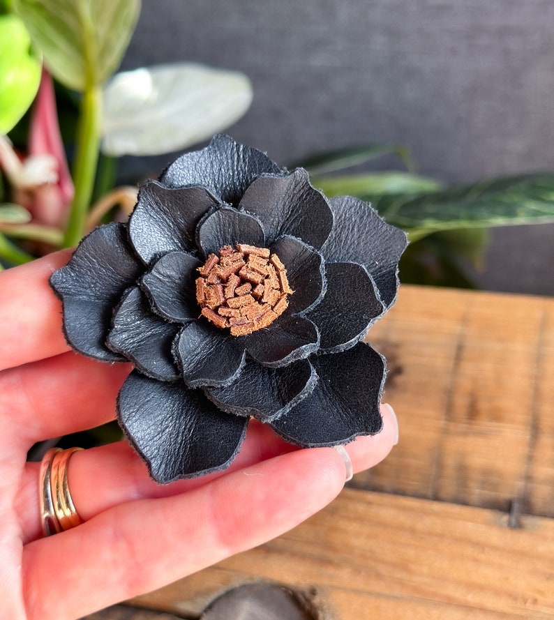 Pode incluir: Uma flor de couro preto com um centro marrom. A flor &eacute; feita de v&aacute;rias camadas de couro e tem uma apar&ecirc;ncia realista.