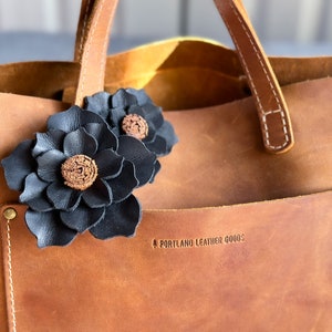 Pode incluir: Uma bolsa tote de couro marrom com detalhes de flores de couro preto. A bolsa tem um bolso na frente com o texto "PORTLAND LEATHER GOODS" em relevo.