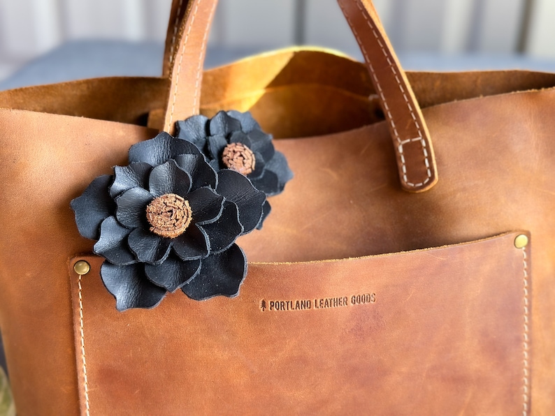 Pode incluir: Uma bolsa tote de couro marrom com detalhes de flores de couro preto. A bolsa tem um bolso na frente com o texto "PORTLAND LEATHER GOODS" em relevo.