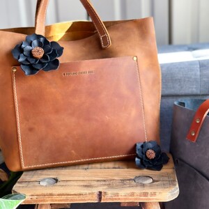 Pode incluir: Uma bolsa tote de couro marrom com um bolso frontal e uma flor de couro preto. A bolsa tem o texto "Portland Leather Goods" gravado no bolso frontal.