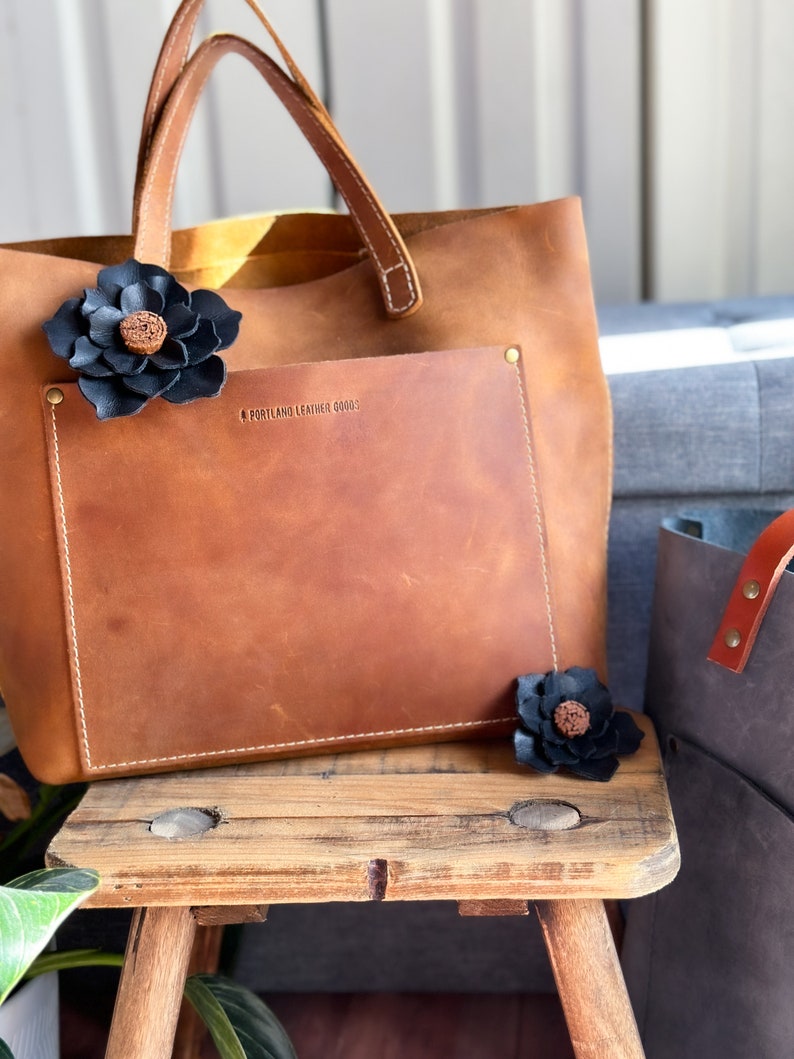 Pode incluir: Uma bolsa tote de couro marrom com um bolso frontal e uma flor de couro preto. A bolsa tem o texto "Portland Leather Goods" gravado no bolso frontal.