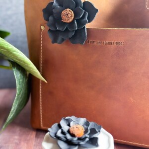 Pode incluir: Uma bolsa tote de couro marrom com um charme de flor de couro preto preso &agrave; al&ccedil;a. A bolsa tem o texto "Portland Leather Goods" gravado na frente.
