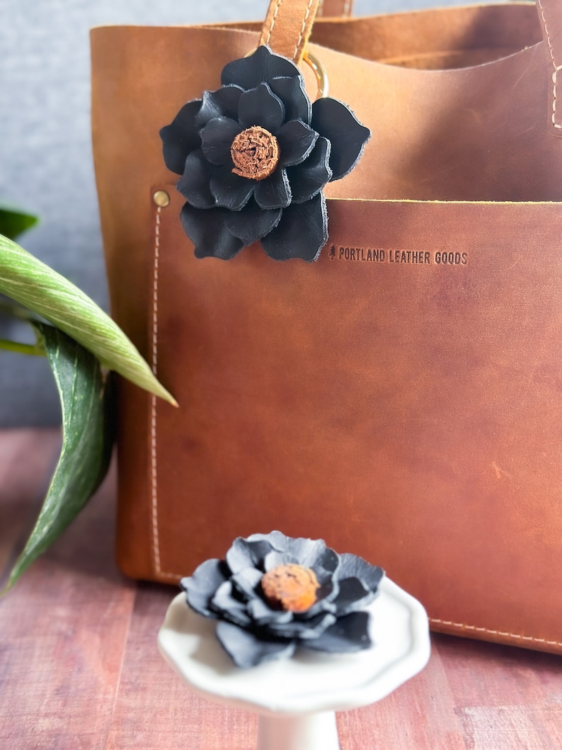Pode incluir: Uma bolsa tote de couro marrom com um charme de flor de couro preto preso &agrave; al&ccedil;a. A bolsa tem o texto "Portland Leather Goods" gravado na frente.