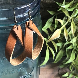 May include: A pair of brown leather earrings with a simple, modern design. The earrings are made of a single strip of leather that is folded and stitched to create a loop. The earrings are hanging from gold-toned hooks.