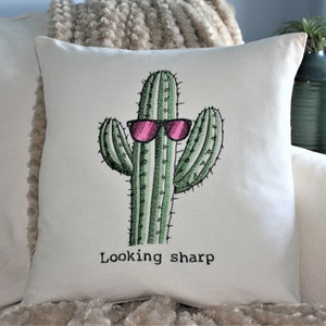 May include: A white pillow with an embroidered cactus wearing pink sunglasses and the text "Looking sharp".