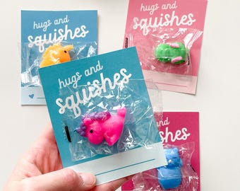 Pode incluir: Quatro cartões coloridos com o texto "hugs and squishes" impresso neles. Cada cartão tem um pequeno brinquedo de plástico em um saco de plástico transparente preso a ele. Os brinquedos são um dinossauro amarelo, um sapo verde, um unicórnio rosa e um gato azul.