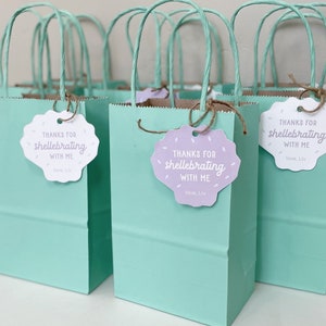 May include: A set of light blue favor bags with white and purple tags that say "Thanks for shellebrating with me, love, Liv".