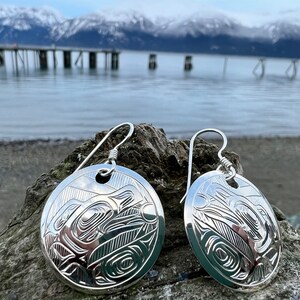 May include: A pair of silver earrings with a circular design featuring a stylized animal, possibly a bear or a wolf, with intricate details. The earrings are hanging from a simple hook.