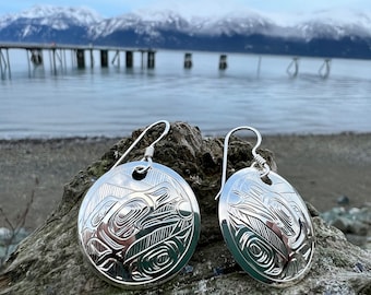 May include: A pair of silver earrings with a circular design featuring a stylized animal, possibly a bear or a wolf, with intricate details. The earrings are hanging from a simple hook.