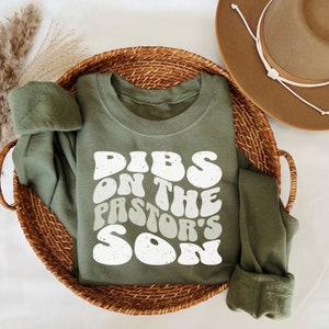 May include: A green long-sleeved sweatshirt with the text "DIBS ON THE PASTOR'S SON" in white, retro-style lettering.