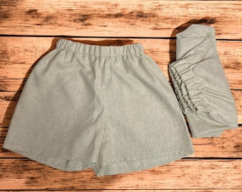 May include: A pair of light grey linen shorts with an elastic waistband. The shorts are folded in half and laid on a wooden surface.