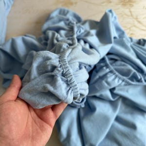 May include: A close-up of a light blue fabric with a gathered elastic band. The fabric is soft and wrinkled, showing the texture of the material.