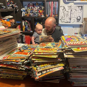 May include: A collection of vintage comic books, including titles like "Conan" and "Marvel Comics Group," stacked on a desk. A man and a baby are looking at a comic book. Shelves in the background are filled with toys and more comics.