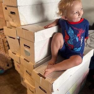 May include: A young child wearing a blue Spider-Man pajama set sits atop a stack of white cardboard storage boxes. The boxes are slightly worn, with some having colorful drawings on them. The child has blonde hair and is looking off to the side.