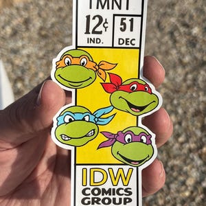 May include: A rectangular sticker featuring the Teenage Mutant Ninja Turtles. The sticker has a yellow background with the four turtles' faces, each wearing a different colored mask. The top of the sticker reads "TMNT" and "12¢ 51". The bottom reads "IDW COMICS GROUP".