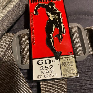May include: A rectangular Marvel pin featuring Spider-Man in a black suit against a red background. The pin includes the Marvel logo and comic details, with the text "60¢ 252 MAY". The pin is attached to a gray backpack strap.