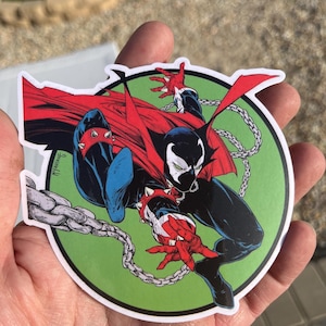 May include: A round sticker featuring a comic book illustration of Spawn. The character is depicted in a dynamic pose with a red cape, black suit, and white mask, set against a green background with a silver chain.