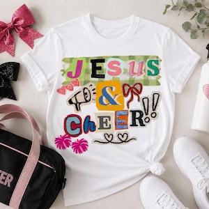 May include: A white t-shirt featuring a colourful graphic with the text JESUS & CHEER, a megaphone, bows, and pom-poms. The design uses various fonts and colours including green, pink, blue, and orange. The shirt is surrounded by cheer accessories.