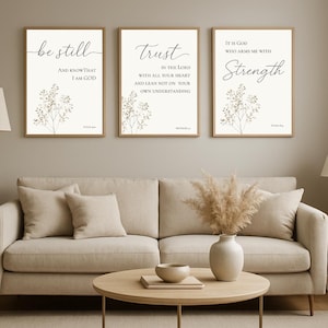 May include: Three framed art prints with inspirational quotes. The prints feature the phrases "be still", "trust", and "Strength" in elegant script, with floral accents. The frames are a warm brown, and the background is a soft cream color.