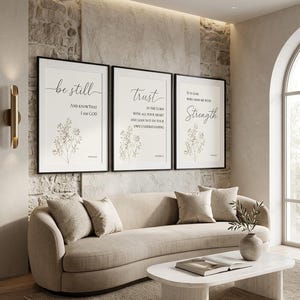 May include: Three framed prints with inspirational quotes: "Be Still," "Trust," and "Strength." Each print features elegant script and botanical illustrations in a neutral colour palette. The artwork hangs above a beige sofa in a modern living room.