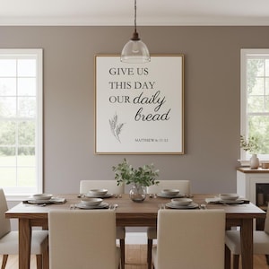 May include: A dining room scene featuring a wooden table set for a meal. Above the table hangs a framed print with the text "GIVE US THIS DAY OUR daily bread" and "MATTHEW 6: 11-13". The room has neutral tones and natural light.