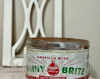 Puede incluir: Una lata de adornos navideños Shiny Brite vintage. La lata cilíndrica tiene una tapa de metal oxidado y una etiqueta con un diseño de adorno rojo y blanco. El texto "AMERICAN MADE" y "GLASS CHRISTMAS TREE ORNAMENTS" son visibles.