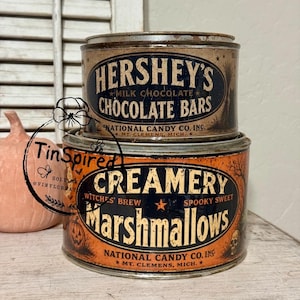May include: Two stacked vintage-style metal tins. The top tin is tan with blue text reading HERSHEY'S MILK CHOCOLATE BARS. The bottom, larger tin is orange with black text reading CREAMERY MARSHMALLOWS, WITCHES' BREW, and SPOOKY SWEET.