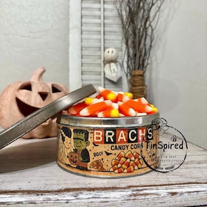 Puede incluir: Una lata redonda de metal Brach's Candy Corn con tapa extraíble, con ilustraciones del monstruo de Frankenstein y una novia, llena de dulces de maíz naranjas, amarillos y blancos. La lata muestra el texto Brach's Candy Corn y Boo!