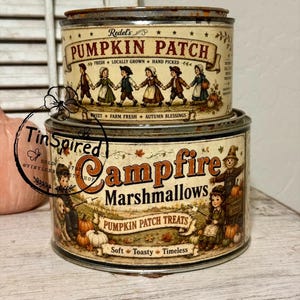 May include: Two stacked cylindrical metal tins with vintage-style graphics. The top tin features children holding hands and the text Redel's PUMPKIN PATCH. The bottom tin displays a campfire scene with the text Campfire Marshmallows PUMPKIN PATCH TREATS.