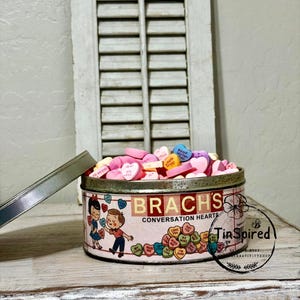 Puede incluir: Una lata vintage de Brach's Conversation Hearts llena de caramelos coloridos en forma de corazón. La lata tiene un fondo rosa con ilustraciones de niños y corazones. La tapa está abierta, revelando los caramelos en el interior.