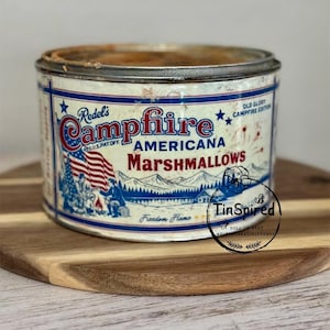 Puede incluir: Lata vintage de malvaviscos Campfire Americana con un diseño patriótico. La lata es blanca con detalles en azul y rojo, y presenta la bandera estadounidense y un paisaje montañoso. Se muestra el texto "Campfire Americana Marshmallows".