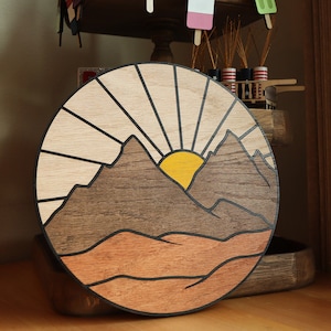 May include: A wooden wall art piece depicting a stylized mountain range with a yellow sun rising behind it. The sun's rays are represented by thin lines radiating outward from the sun. The mountain range is brown and the background is a light wood color.